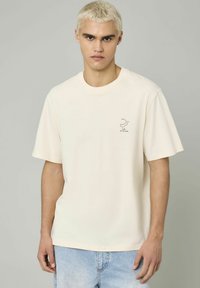 T-shirt en coton crème avec des manches courtes, un col rond et un petit logo graphique sur la poitrine gauche. Coupe décontractée avec une texture lisse.