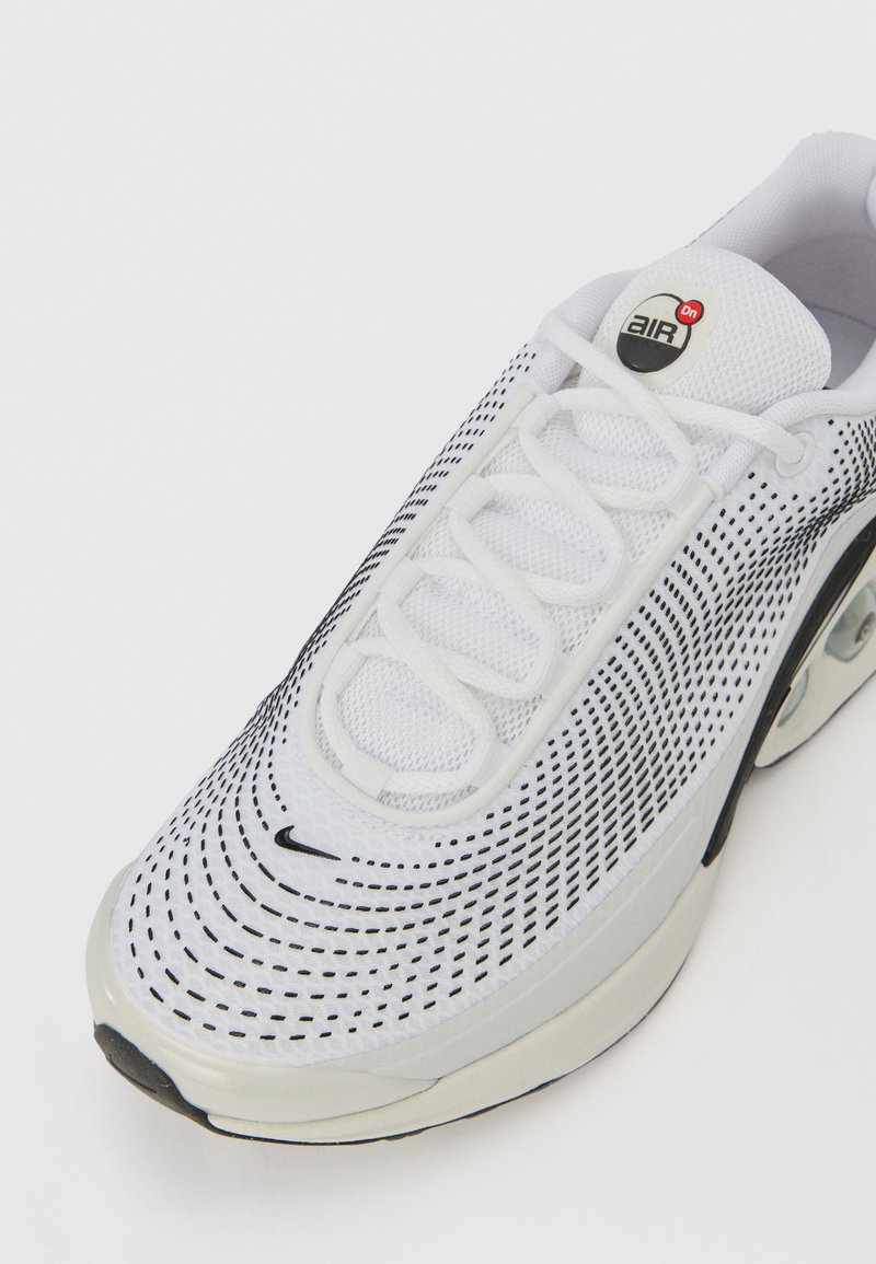 Chaussure de sport blanche avec une tige en mesh ornée de motifs noirs en points, des lacets ronds et une unité de coussin d'air visible dans la semelle.