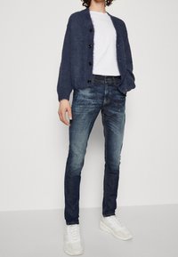 Cárdigan de punto azul marino con cierre de botones sobre una camiseta blanca, combinado con jeans azul claros y zapatillas blancas. Presentación de un atuendo casual.