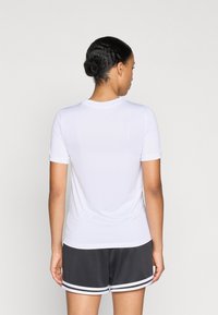 Cămașă sport albă cu mâneci scurte, guler rotund, textură netedă și siluetă ajustată, purtată cu pantaloni scurți închiși la culoare cu dungi albe.