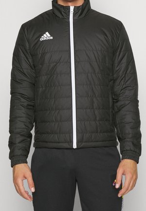 Mann trägt eine schwarze Adidas-Steppjacke mit weißem Reißverschluss und Adidas-Logo auf der Brust, kombiniert mit schwarzen Hosen.