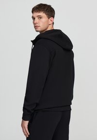 Giovane uomo in felpa nera e pantaloni sta in profilo, guardando oltre la spalla contro uno sfondo grigio chiaro uniforme.