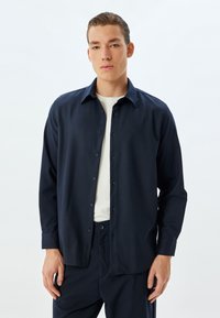 Marineblauw overhemd met lange mouwen, een klassieke kraag en een gladde textuur. Gekozen boven een witte ronde hals, gecombineerd met bijpassende broek.