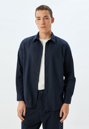 Camisa azul marino de botones con mangas largas, un cuello clásico y una textura suave. Llevada sobre un suéter blanco de cuello redondo, combinada con pantalones a juego.