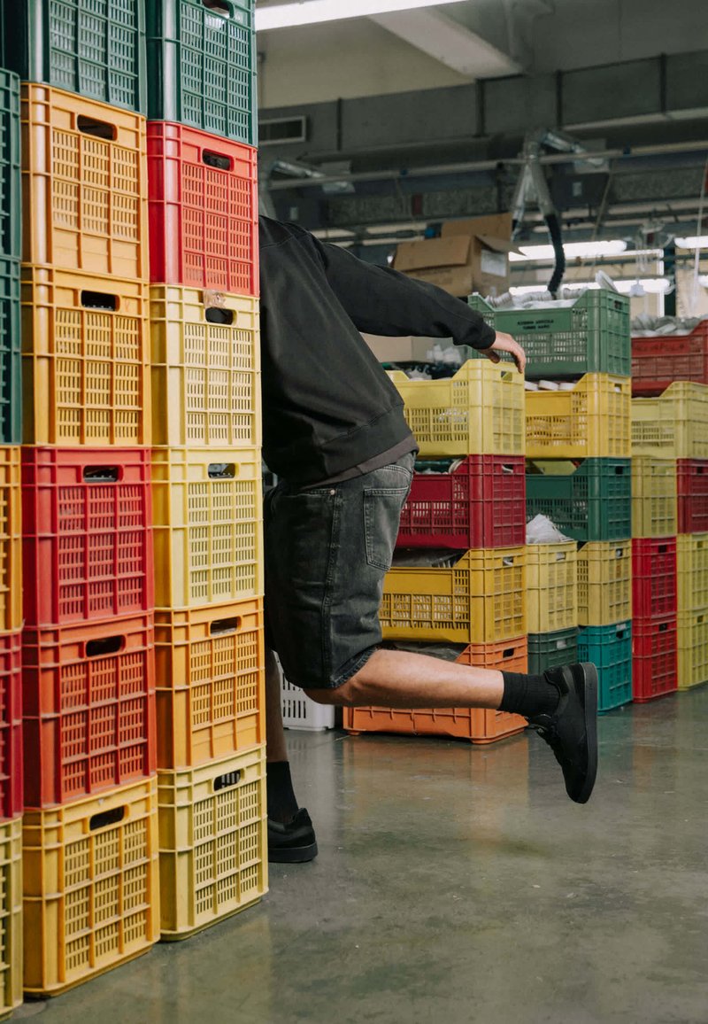 Personne vêtue de noir en équilibre sur une jambe parmi de hautes piles de caisses en plastique rouges, jaunes, vertes et orange dans un environnement industriel.