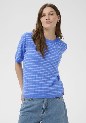 Młoda kobieta z długimi brązowymi włosami, ubrana w niebieski sweter w kratę z krótkim rękawem i jasnoniebieskie dżinsy, stojąca na jednolitym tle.