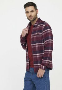 Chemise en flanelle à carreaux rouges et bleu marine avec des manches longues et une poche poitrine, superposée à un T-shirt bordeaux. Jean à coupe classique.
