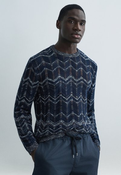 Navy blue knit sweater with a geometric herringbone pattern, featuring a crew neckline and contrasting textured trim. Worn with gray drawstring pants.