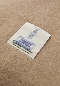 Brun filtstoff med et vevd merke som har en lyseblå illustrasjon av et monument og svart tekst som står "Originals Studio."