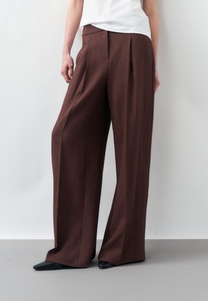 Personne portant un pantalon large marron, un t-shirt blanc et des chaussures noires pointues, debout contre un fond clair uni.