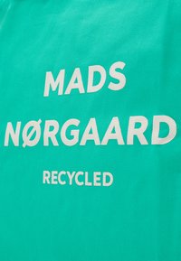 Türkiz szövet, amelyen bátor, fehér betűk olvashatók: "MADS NØRGAARD" és "RECYCLED" egy egyszerű, sans-serif betűtípussal.