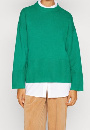 Pull en tricot vert avec un col haut, ourlet côtelé et épaules tombantes, superposé à une chemise blanche à col, associé à un pantalon en velours côtelé beige.
