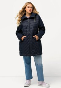 Cappotto imbottito blu navy con chiusura a zip, design trapuntato nella parte superiore, doppie tasche laterali e cappuccio a contrasto. Indossato con jeans azzurri chiari e sneakers.