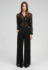 Femme aux cheveux longs portant une chemise à boutons en dentelle noire et un pantalon large en dentelle assorti, se tenant avec une main dans la poche.