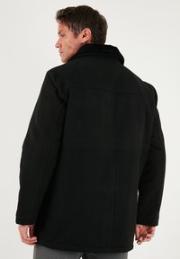 Melns vilnas mētelis ar teksturētu apdari, plašu apkakli un strukturētām plecu daļām, ar piegriezumu, kas akcentē figūru, un redzamiem šuvējiem. 