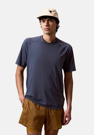 Jonge man met een beige pet, een donkergrijs kortärmelig shirt en een bruine korte broek, staand met één hand in zijn zak tegen een effen achtergrond.