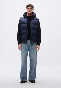 Colete acolchoado azul-marinho com capuz, apresentando um design acolchoado. Usado sobre um suéter preto texturizado e combinado com jeans azul claro de perna larga.