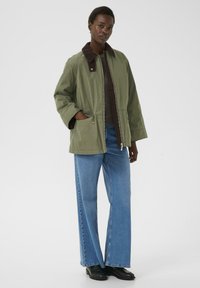 Jachetă verde măslinie cu guler din fustă, fermoar frontal și două buzunare laterale. Asortată cu blugi din denim albastri cu picioare largi și botine negre.