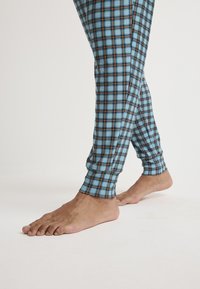 Blå rutig pyjamasbyxor har ett rutmönster med svarta och bruna accenter. Bensluten är tighta och tyget verkar mjukt. Fötterna är barfota.