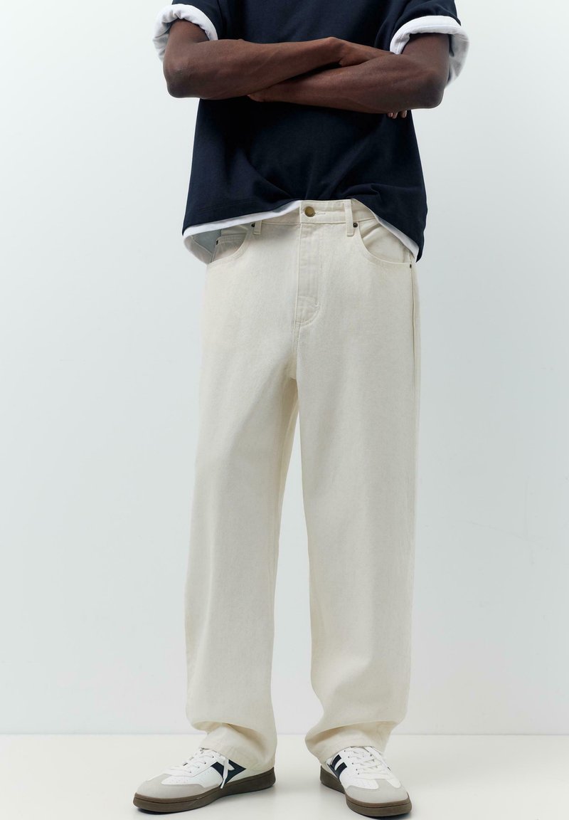 Homme portant un pantalon ample couleur crème, des baskets blanches et noires, et une chemise noire à manches longues retroussées, les bras croisés.