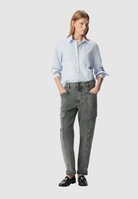 Camicia a righe blu e bianche con maniche lunghe, abbinata a pantaloni cargo grigi con grandi tasche e scarpe nere.