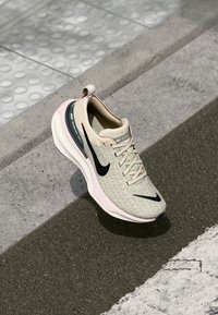 Beige Sport Schuh mit strukturiertem Obermaterial, schwarzem Swoosh, rosa gefärbter Sohle und minimalistischem Design. Mit einer Schlaufe an der Rückseite.