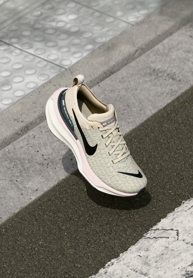 Beige Sport Schuh mit strukturiertem Obermaterial, schwarzem Swoosh, rosa gefärbter Sohle und minimalistischem Design. Mit einer Schlaufe an der Rückseite.