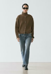 Brauner gerippter Strickcardigan mit hohem Kragen, Knopfleiste und überschnittenen Schultern, kombiniert mit hellblauen Flared-Jeans und schwarzen Stiefeletten.