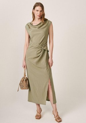 Mujer con vestido midi sin mangas de color verde oliva con detalle de nudo y abertura lateral, sosteniendo un bolso beige y llevando sandalias de tacón color marrón claro.