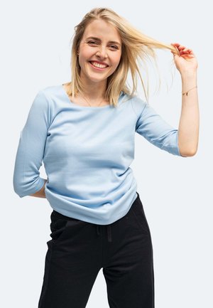 Junge Frau lächelt, spielt mit ihrem blonden Haar, trägt ein hellblaues Langarmshirt und schwarze Hose vor einem einfarbigen Hintergrund.