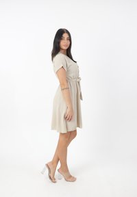 Vestito beige a maniche corte con bottone frontale e vita legata. Presenta una gonna fluente e sandali con tacco trasparente. Tessuto semplice e texture.