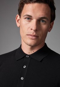 Sort poloshirt med tekstureret strikfinish, tre-knaps knaplukning og klassisk krave. Modellens overkrop er synlig mod en neutral baggrund.