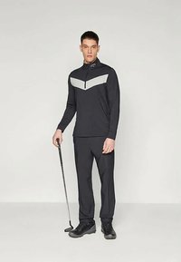 Svart langermet zippered topp med grå chevron-design, akkompagnert av svarte treningsbukser og sorte sko. Modell holder en golfkølle.