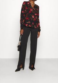 Schwarze Blumenshirt mit roten und grünen Mustern, kombiniert mit schwarzen Hosen mit Seitenschlitzen und spitzen schwarzen Absätzen. Kleine schwarze Handtasche.