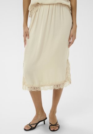 Beżowa spódnica midi z koronkową lamówką na dole i po bokach, noszona z czarnymi sandałami na obcasie z paskami, widoczna od talii do stóp.