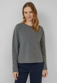 Femme aux cheveux châtain clair portant un pull gris à manches longues et un pantalon bleu foncé, se tenant devant un fond gris clair uni.
