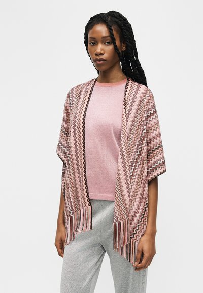 Femme aux longs cheveux tressés portant un châle rose à motif zigzag sur un haut rose et un pantalon gris clair, devant un fond uni.