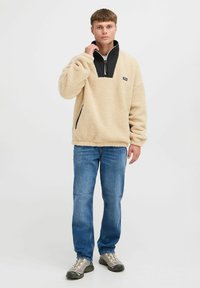 Beige fleece pullover with a black collar and a quarter-zip, featuring a small logo on the chest and side pockets. Worn with blue jeans.