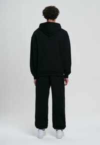 Felpa nera con cappuccio e coulisse, vestibilità ampia e polsini a costine, abbinata a pantaloni della tuta neri e sneakers bianche.