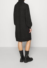Abito maglione nero a maglia con collo alto, maniche a palloncino e vestibilità ampia. Materiale strutturato e orli a coste. Indossato con stivali neri.