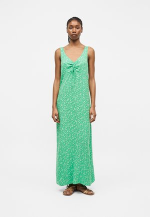 Femme debout portant une robe maxi verte sans manches avec un imprimé floral blanc et un froncé à l'avant, associée à des sandales plates marron à brides.