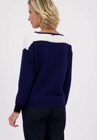 Vrouw met kort blond haar, gekleed in een marineblauwe trui met een witte strook over de bovenrug, gecombineerd met donkere spijkerbroeken, met haar gezicht naar links.