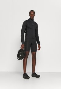 Czarna kurtka rowerowa z wysokim kołnierzem, pełnym zamkiem i dopasowanym krojem; zestawiona z czarnymi spodenki z poduszkami i dopasowanym kaskiem.