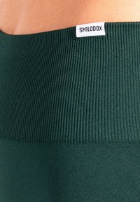 Groen geribd materiaal met een gladde textuur. Voorzien van een logolabel "SMILODOX" dat aan de tailleband is bevestigd. Het ontwerp legt de nadruk op comfort en pasvorm.