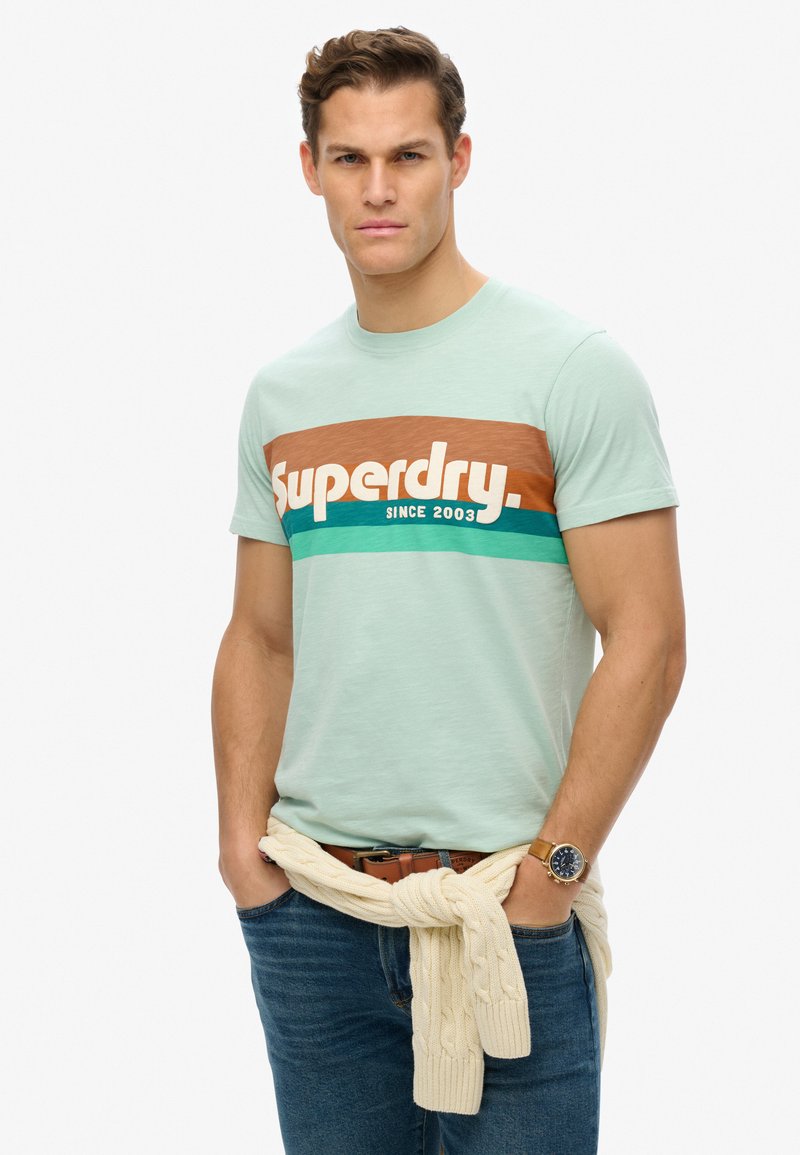 Light green cotton t-shirt with a brown, teal, and white graphic design. Model wears it paired with dark jeans and a cream sweater tied at the waist.