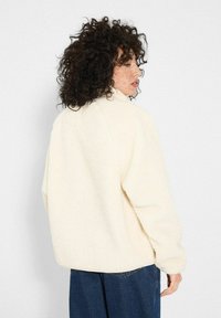 Kobieta z kręconymi włosami, ubrana w kremowy polar i niebieskie dżinsy, patrzy w bok na jasnoszarym tle.