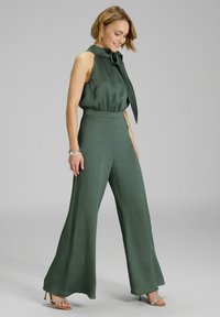 Groene wijde jumpsuit met een mouwloos ontwerp, een striknek, gestructureerde stof en een getailleerde taille, gecombineerd met sandalen met hak en bandjes.