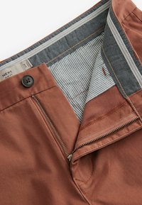 Pantalon slim fit en tissu orange brûlé, avec une ceinture rayée contrastante. Il dispose d'une fermeture éclair avant et d'un bouton pour un maintien sécurisé.