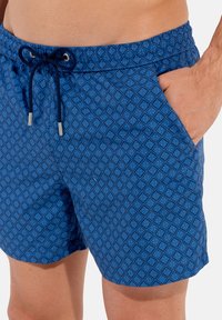 Shorts de bain bleus avec un motif géométrique en losanges, taille élastique, fermeture à cordon de serrage et poches latérales. Texture lisse.