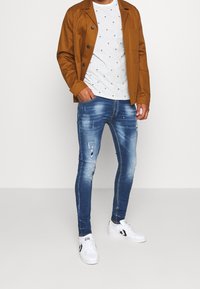 Man som bär en brun knäppt jacka, en vit randig t-shirt med små svarta palmträd, slitna blå skinny jeans och vita sneakers.
